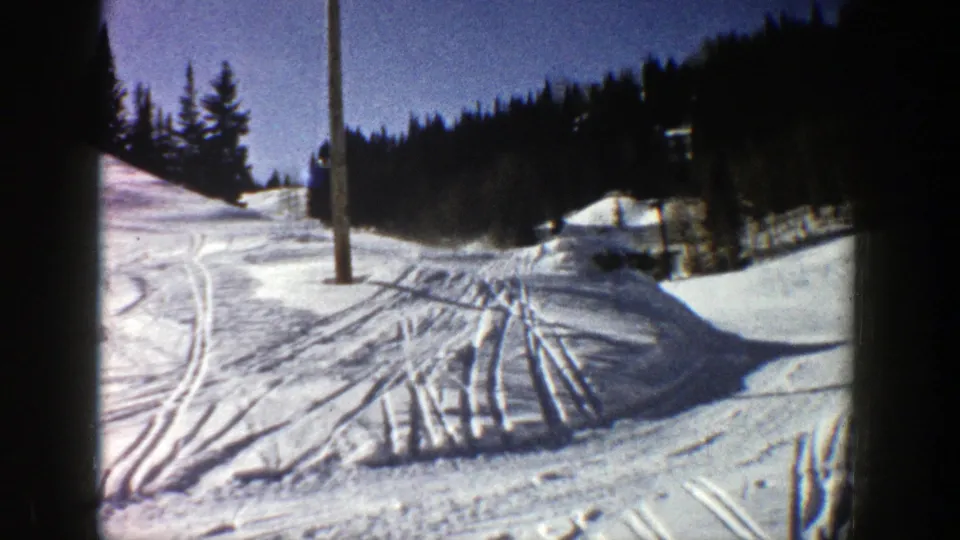 Home-movie view of an Aspen ski run with multiple tracks, a lift or utility pole, evergreens, and bright winter light.