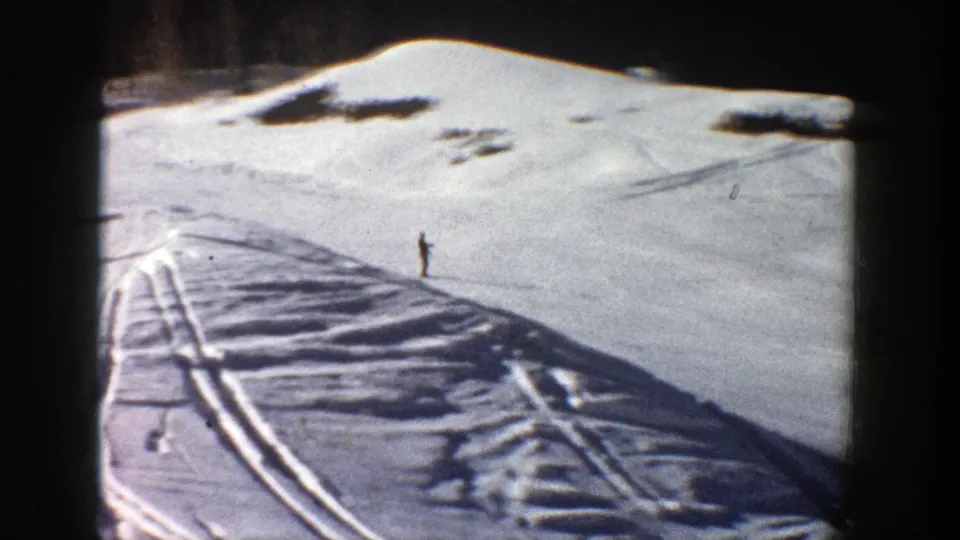 1961 Aspen frame with a distant skier low on the slope, wide snowfield, ski tracks, and dark tree line at the top.