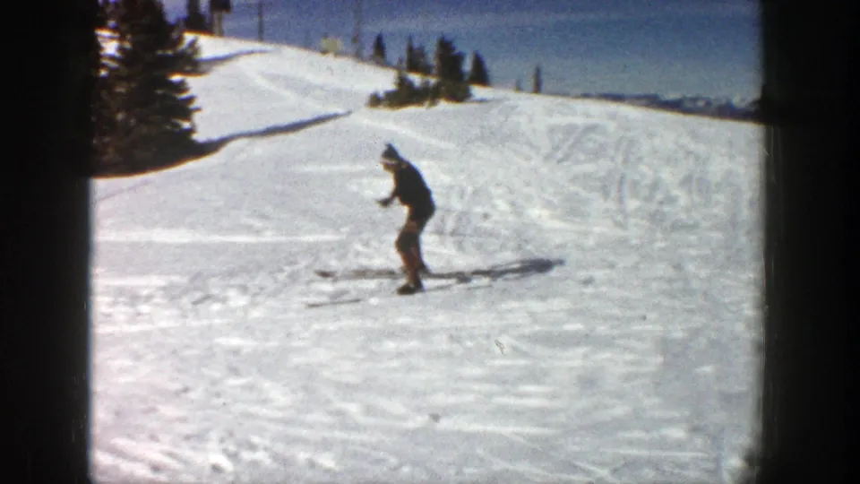 A lone skier in dark winter clothing crosses a snow-covered Aspen slope in 1961 home-movie footage, with evergreen trees behind.