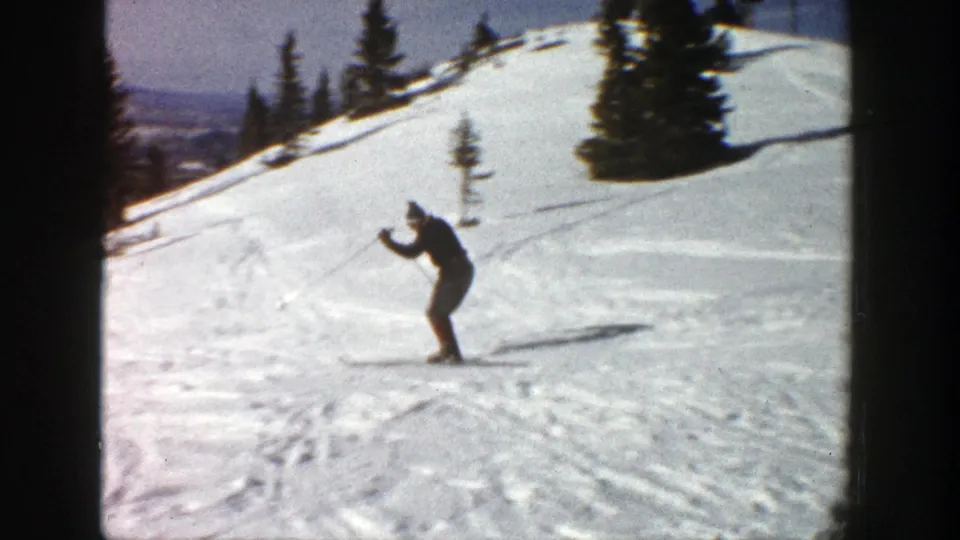 A skier on an Aspen snow slope appears to steady with poles while evergreen trees line the upper hillside in this 1961 8mm frame.
