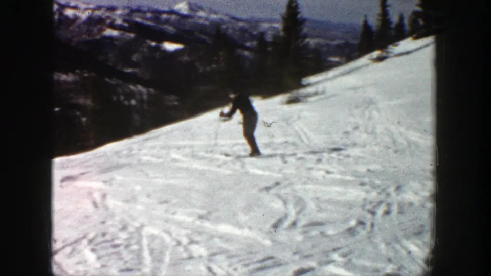 A distant skier descends an open Aspen slope in 1961, with snow tracks, evergreens and mountain ridges visible beyond the run.