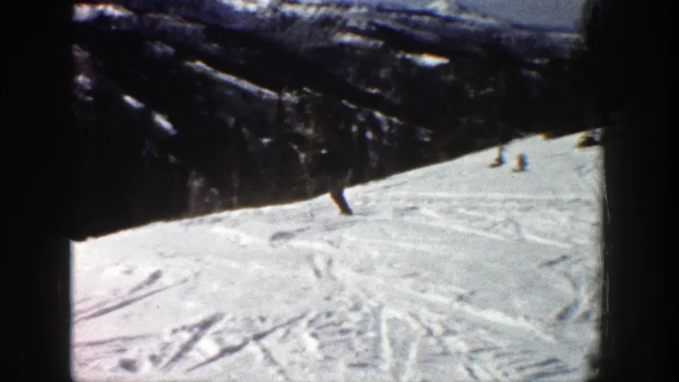 A small figure skis across a snowy Aspen hillside in 1961 footage, with dark forested mountain terrain and tracked snow in view.