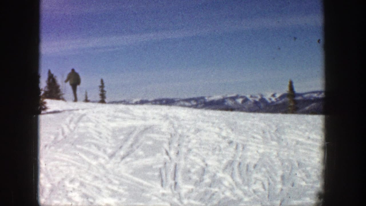 Distant skier walking on a snowy slope with tracks, evergreens, mountains, and blue sky.
