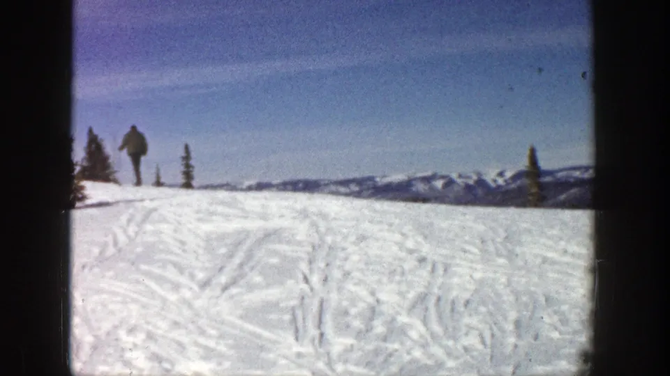 In 1961 Aspen, a distant skier in dark clothing walks uphill across a tracked snowy slope with evergreens and mountain ridges beyond.