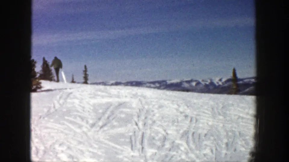 A distant skier continues uphill on an Aspen snow ridge in 1961, framed by sparse evergreens, tracked snow, and blue mountain horizon.