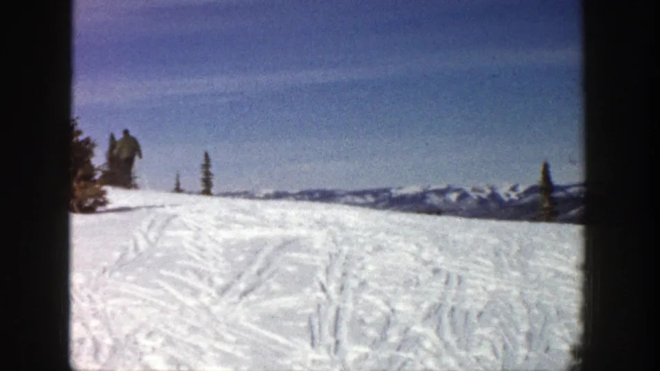The 1961 Aspen home-movie view places the skier small at the left edge of a broad snowfield, with ski tracks and distant peaks visible.