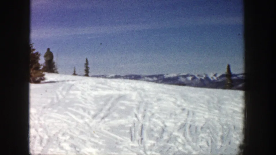 At the end of the 1961 Aspen clip, the skier stands or walks on the snowy hilltop with tracked snow and mountain ridges in the distance.