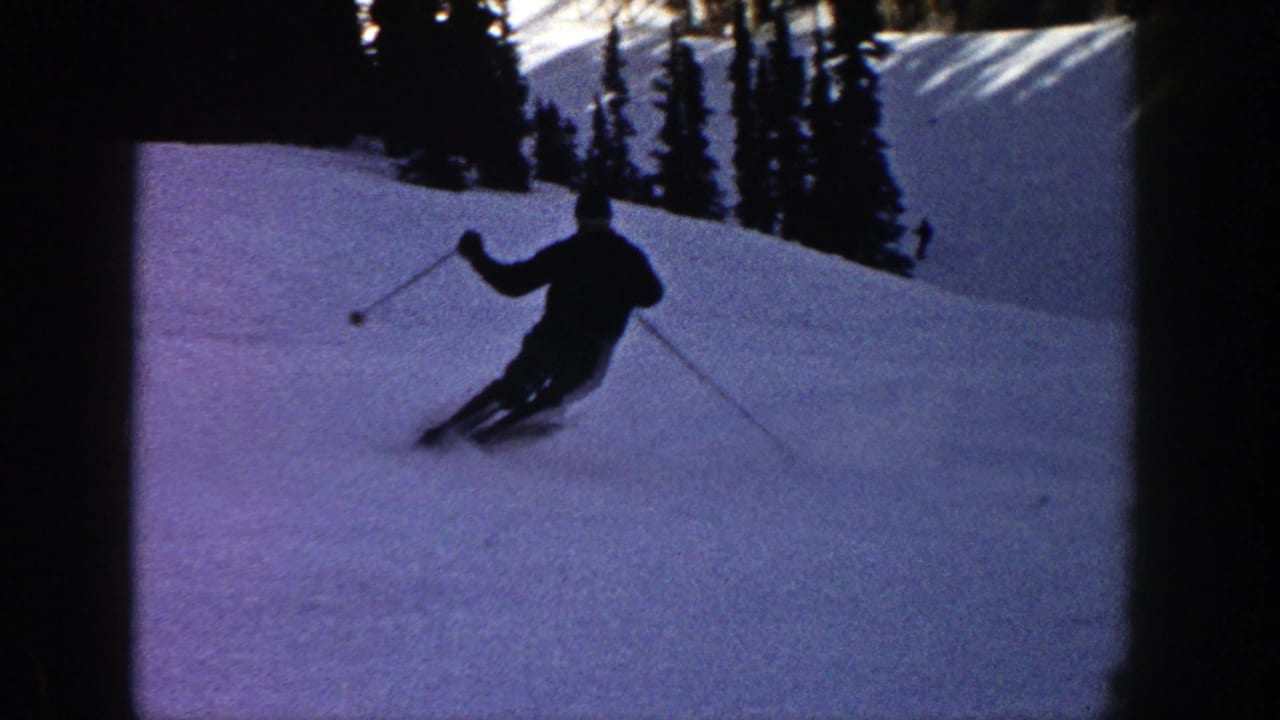 In 1961 Aspen, Colorado footage, a dark-clad skier uses poles while turning on a shaded snowy slope backed by pine trees.