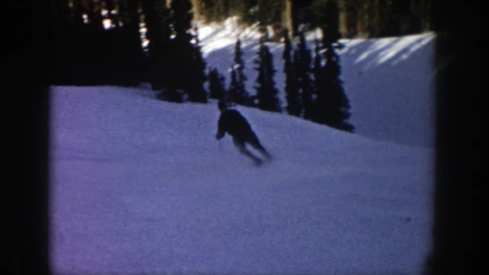 The skier appears farther downhill on the Aspen snowfield, leaning into a turn beneath dark pine trees in low winter light.
