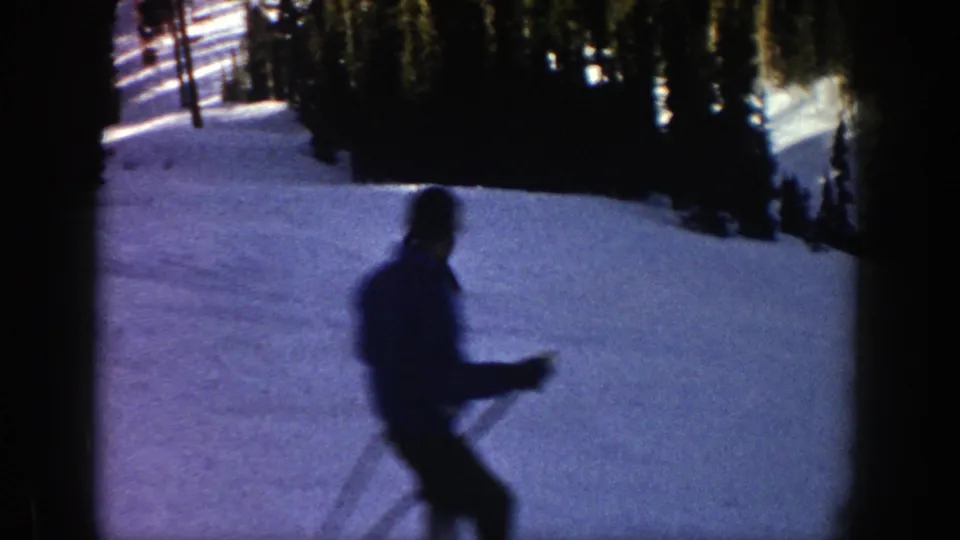 A skier passes closer to the 8mm camera, blurred by motion while holding poles against the blue-toned snow and tree line.