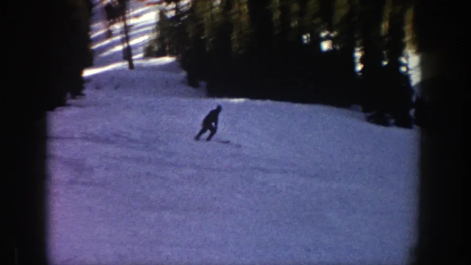 A small silhouetted skier continues down the Aspen slope, framed by shaded snow and dense pines in the background.