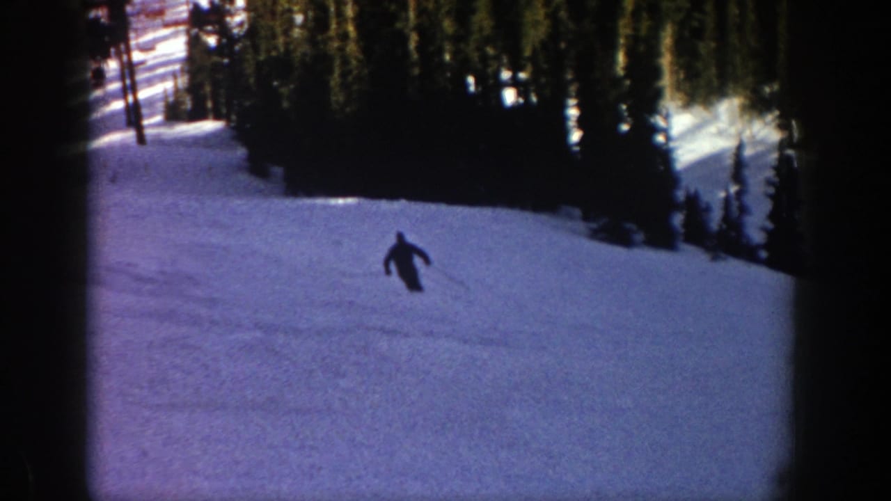 A lone skier in dark clothing moves downhill on a snowy Aspen slope in 1961, framed by tall evergreen trees and 8mm film borders.