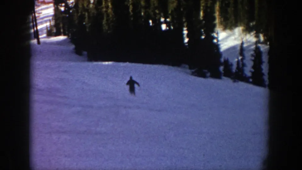 The skier appears farther down the Aspen slope, moving through blue-toned snow with dense evergreen trees across the upper frame.