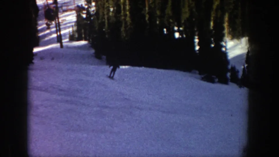 A dark-clad skier passes near the left edge of the 8mm frame on a shaded snowy Aspen run lined with evergreens.