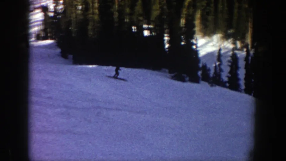 The skier crosses the lower-right portion of the Aspen slope, silhouetted against snow with dense evergreen trees in the background.