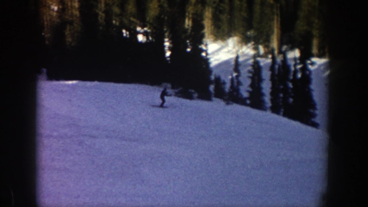 A distant skier moves across a bright snowy Aspen slope in 1961 home-movie footage, with evergreen trees rising behind.