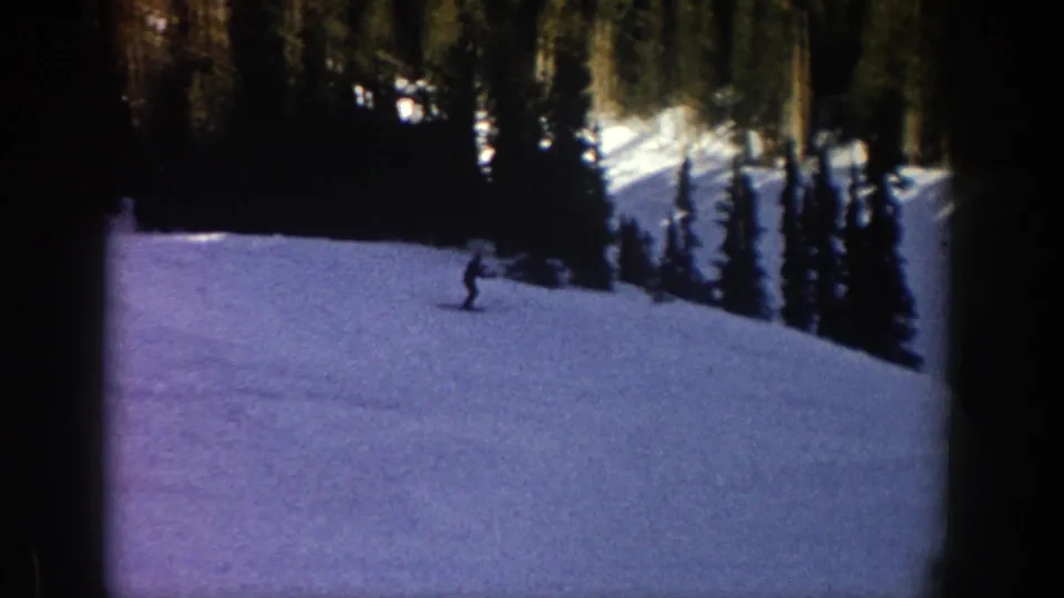 A distant skier moves across a bright snowy Aspen slope in 1961 home-movie footage, with evergreen trees rising behind.
