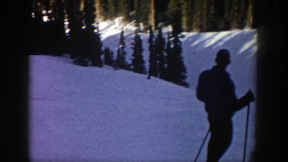 A skier in silhouette stands near the camera on an Aspen snow slope, holding poles against a backdrop of evergreens.