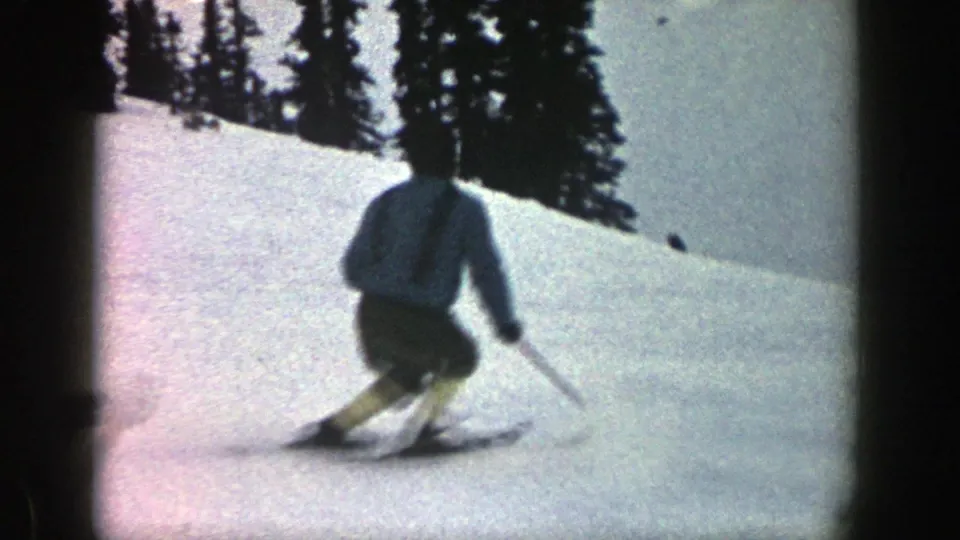 A skier in a light jacket and dark shorts turns downhill on a snowy Aspen slope, filmed in grainy 8mm color.