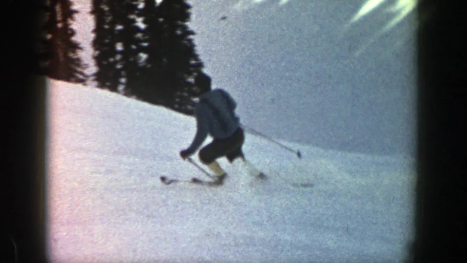 A skier crouches into a sweeping turn on an Aspen snow slope, with ski tracks, poles, and evergreen trees visible.