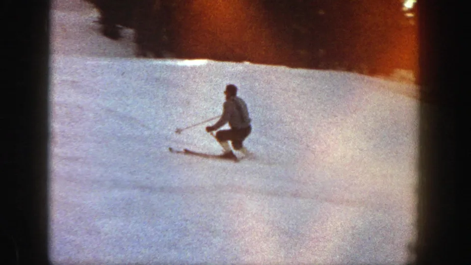 A skier in vintage 8mm Aspen footage crouches into a downhill turn on a bright, open snowfield.
