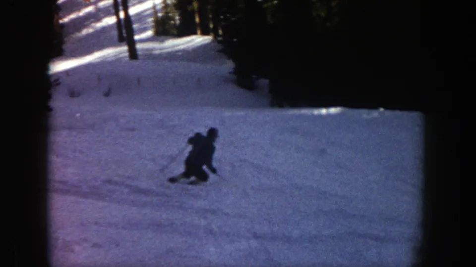 A distant skier moves downhill through shadowed snow in Aspen home-movie footage, with dense evergreens nearby.