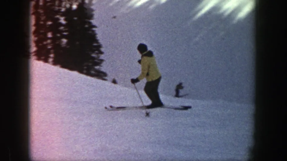 A skier in a yellow jacket descends an Aspen snow slope, with dark evergreen trees along the upper edge.