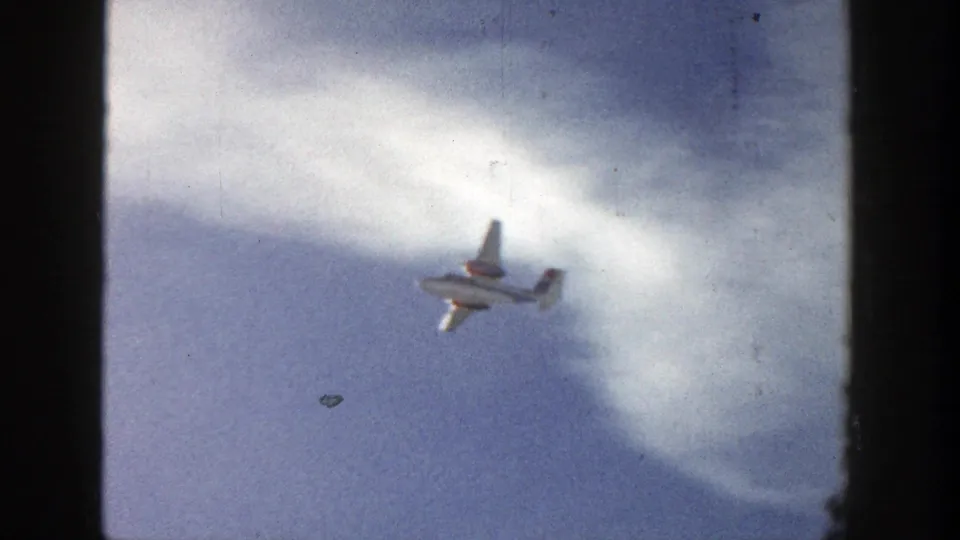 A white aircraft passes beneath a curved cloud band, with grain and dust visible in the 1961 Aspen 8mm footage.