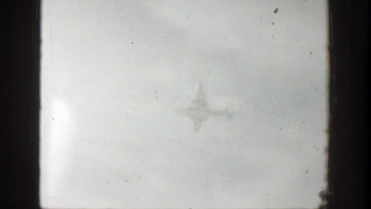 1961 Aspen home-movie frame showing a distant airplane silhouette against a bright, clouded sky.