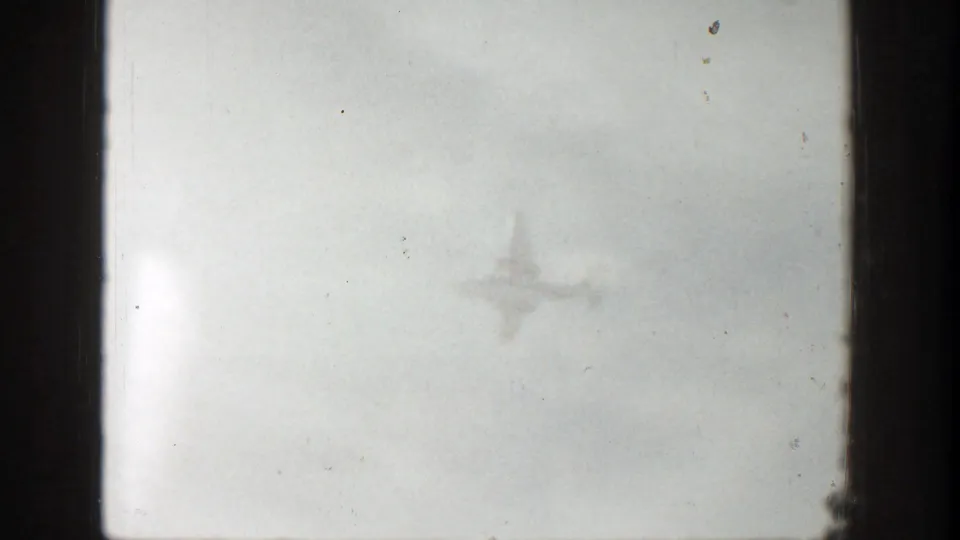 1961 Aspen home-movie frame showing a distant airplane silhouette against a bright, clouded sky.