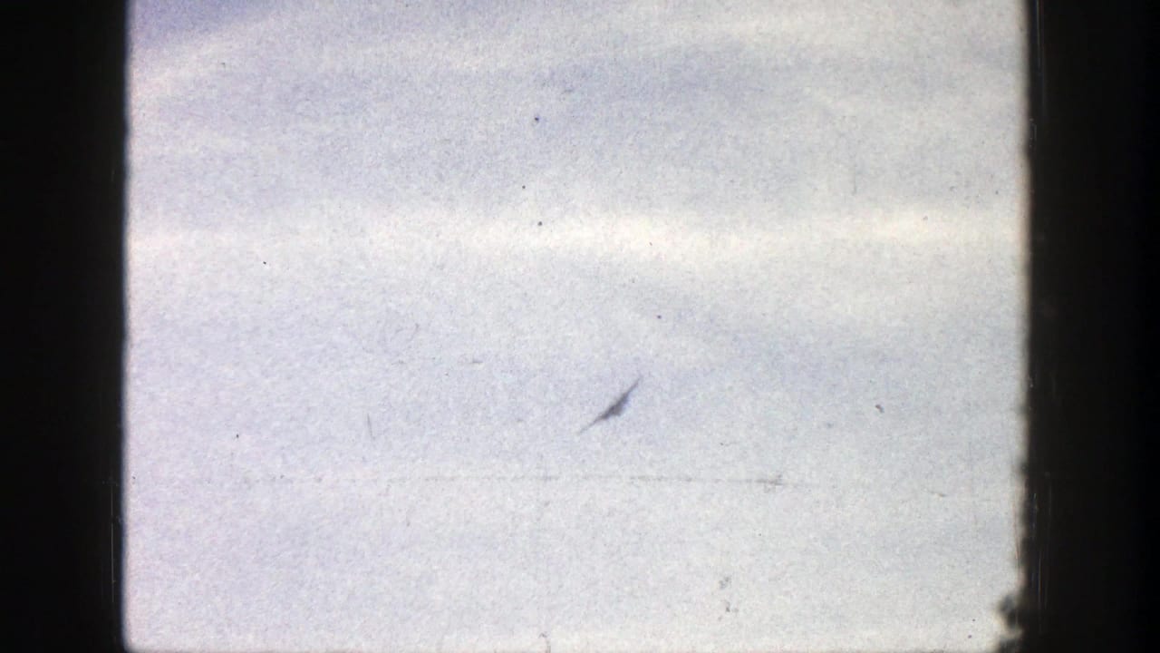 A very distant plane appears against a bright sky in 1961 Aspen, Colorado home-movie footage, with 8mm grain and gate shadow visible.