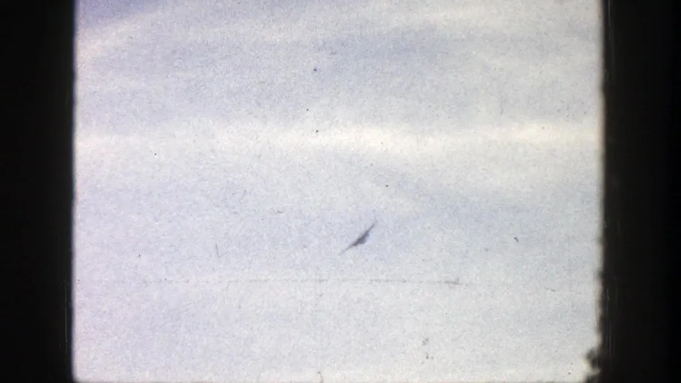 A very distant plane appears against a bright sky in 1961 Aspen, Colorado home-movie footage, with 8mm grain and gate shadow visible.