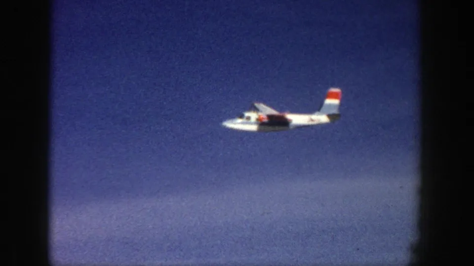 Closer 8mm frame of a white aircraft with red and blue tail markings flying across a deep blue sky.