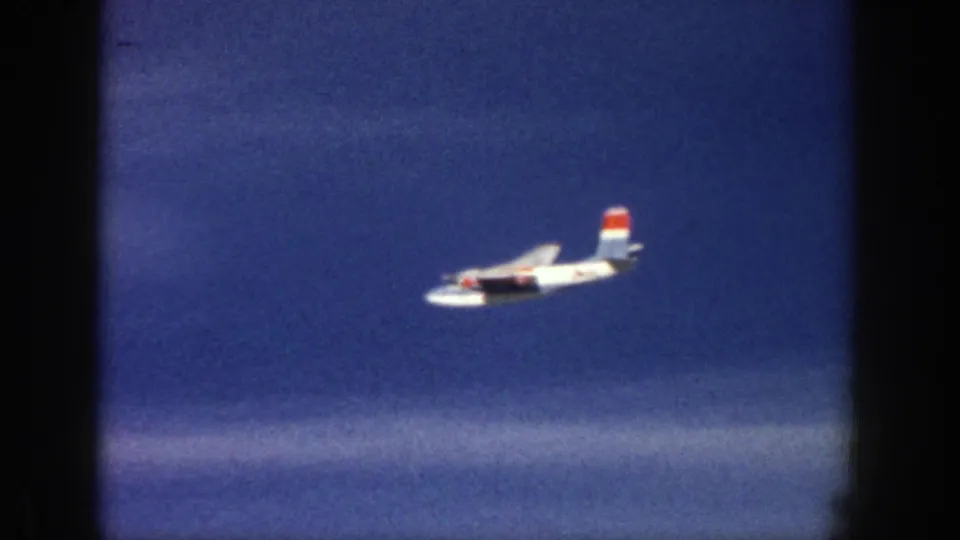 White aircraft with red and blue tail markings crossing a blue sky in the final 1961 Aspen home-movie frame.