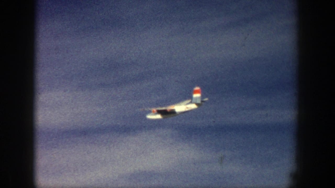 Small propeller airplane crossing a blue sky with dark film borders.