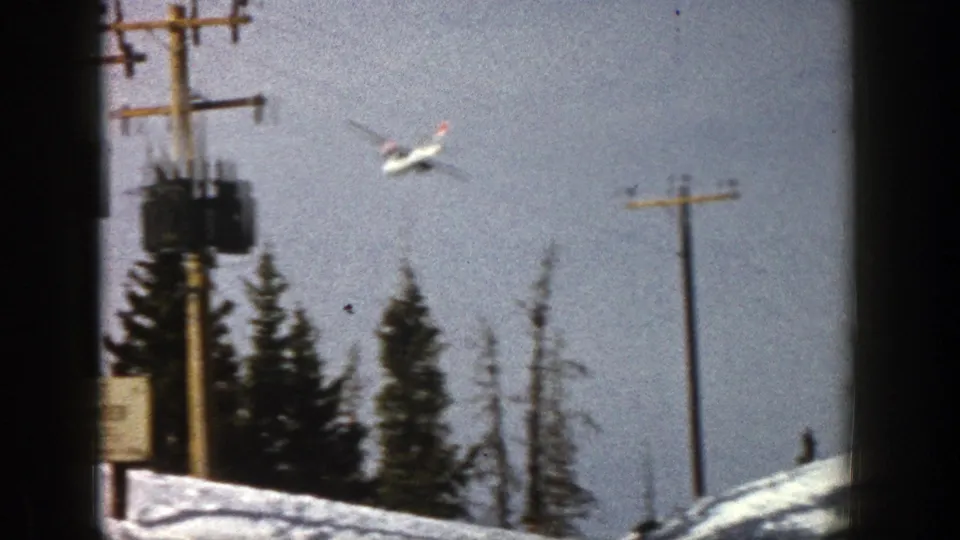 A small aircraft descends over a snowy Aspen-area scene with evergreens, utility poles, and part of a roadside sign visible.