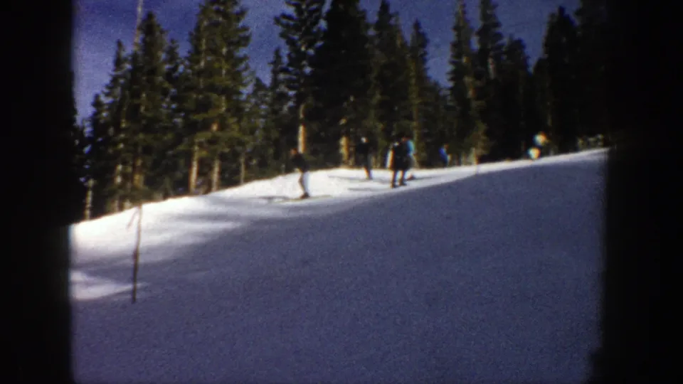 Distant skiers on a snowy Aspen slope in 1961, with evergreen forest and clear sky behind them.