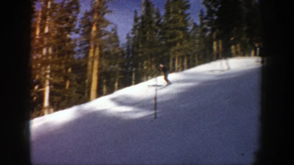 A skier on a 1961 Aspen slope appears to make a small jump or crest over a snow mound among evergreens.