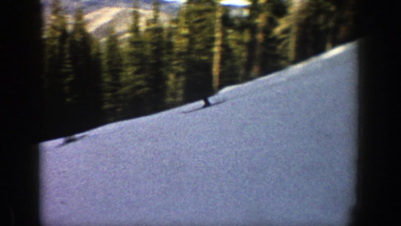Distant skier crossing a snowy forested slope with dark film borders.