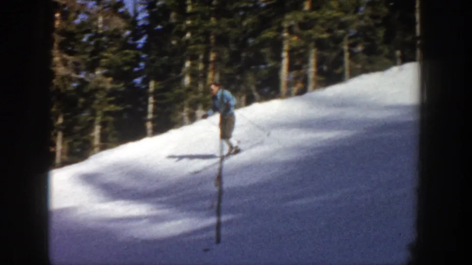 A skier in a blue jacket and dark pants descends an Aspen, Colorado slope in 1961, using poles near a line of evergreen trees.