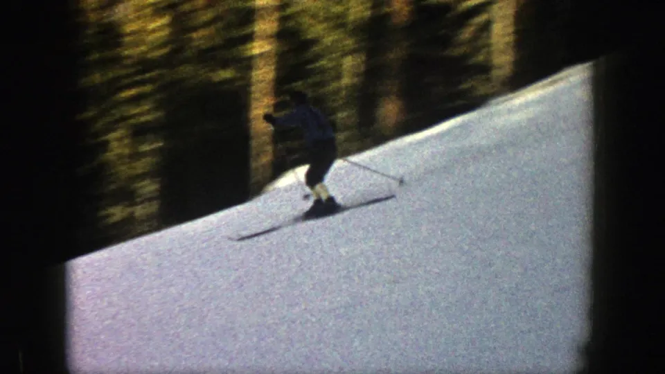 1961 Aspen 8mm frame with a skier crossing a white slope, backed by tall evergreen trees and dark frame edges.