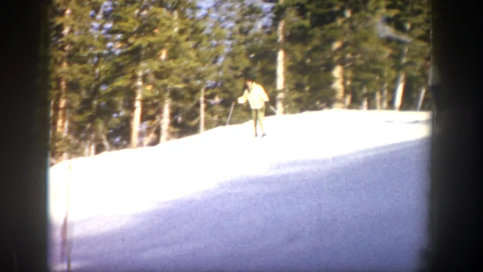1961 Aspen 8mm frame showing a light-jacketed skier on a bright snowfield at the edge of an evergreen forest.