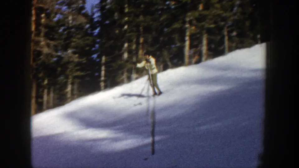 1961 Aspen 8mm frame with a light-jacketed skier in green pants descending a snowy slope before dark evergreens.