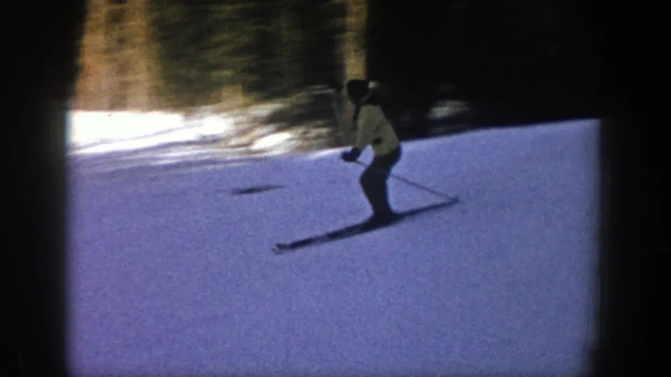 A distant skier in a light jacket bends into the descent on a snowy 1961 Aspen slope, with trees at the edge of frame.