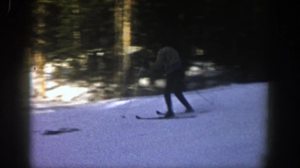 A distant skier in dark pants and a light jacket moves through shadow on the Aspen slope in 1961 home-movie footage.