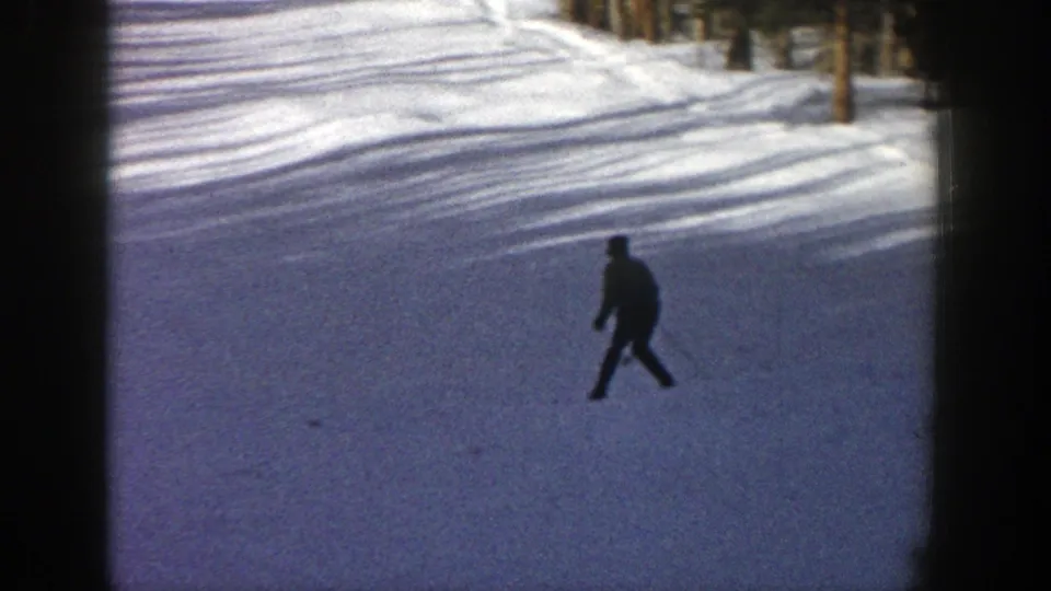 A small skier figure moves across an open Aspen snow slope, framed by trees and long winter shadows.