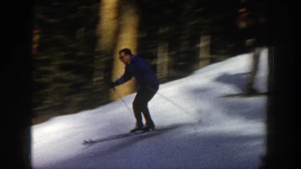 A man in a blue coat skis downhill on a snowy Aspen slope, with trees and strong winter light behind him.