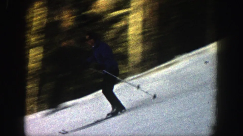 The skier in a blue coat leans across the snowy slope near the tree line, captured with soft 8mm motion blur.