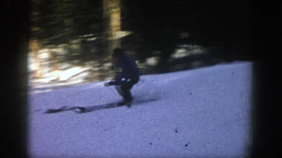 A skier crosses a darker patch of snowy slope in Aspen, with forest shadows and film grain giving the frame a home-movie look.