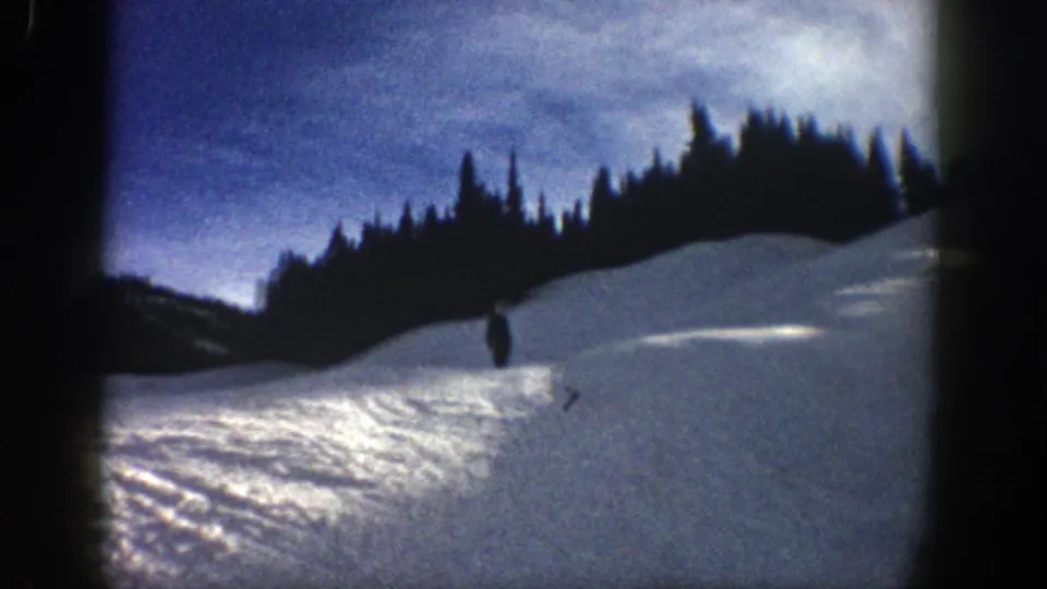 A skier approaches the camera on a snowy Aspen slope, with trees silhouetted against the sky in 1961 home-movie footage.